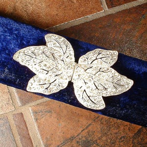 Peut inclure: Une broche argent&eacute;e en forme de feuille avec des accents de cristaux transparents repose sur un tissu de velours bleu fonc&eacute;. La broche pr&eacute;sente des d&eacute;tails complexes, et le tissu a un aspect doux et textur&eacute;.