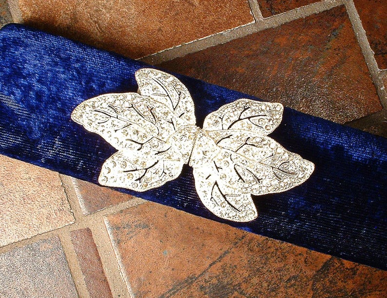 Peut inclure: Une broche argent&eacute;e en forme de feuille avec des accents de cristaux transparents repose sur un tissu de velours bleu fonc&eacute;. La broche pr&eacute;sente des d&eacute;tails complexes, et le tissu a un aspect doux et textur&eacute;.