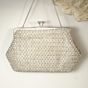 May include: A silver-toned evening clutch bag completely covered in small, clear, reflective rhinestones. The bag features a structured metal frame, a kiss-lock closure with two small decorative knobs, and a thin, detachable silver-toned chain strap.