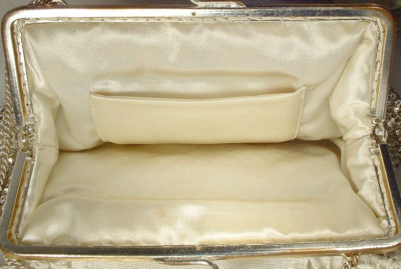 May include: An open rectangular clutch bag featuring a metallic gold-tone frame with a hinged clasp closure. The interior is lined with smooth cream-colored satin fabric and includes a single horizontal slip pocket along the back wall.
