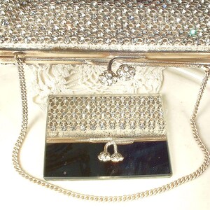 May include: A silver-toned clutch bag covered in clear rhinestones with a metal chain strap and a dual-flower clasp, paired with a matching rectangular compact mirror featuring a rhinestone-encrusted top half and a smooth, reflective bottom half.