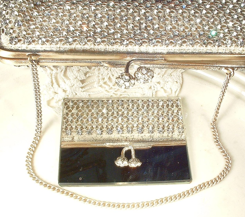 May include: A silver-toned clutch bag covered in clear rhinestones with a metal chain strap and a dual-flower clasp, paired with a matching rectangular compact mirror featuring a rhinestone-encrusted top half and a smooth, reflective bottom half.