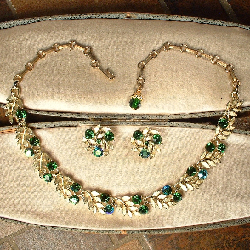 May include: A vintage jewelry set featuring a gold-tone necklace and matching earrings. The necklace and earrings are adorned with green and blue-green rhinestones and leaf-shaped accents. The set is displayed in a vintage jewelry box.