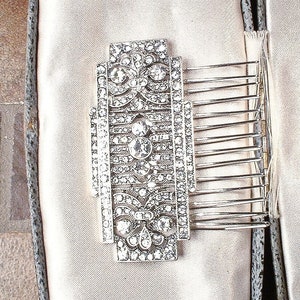 May include: A silver hair comb with a decorative design of clear rhinestones. The comb has a rectangular shape and is set on a white satin background.