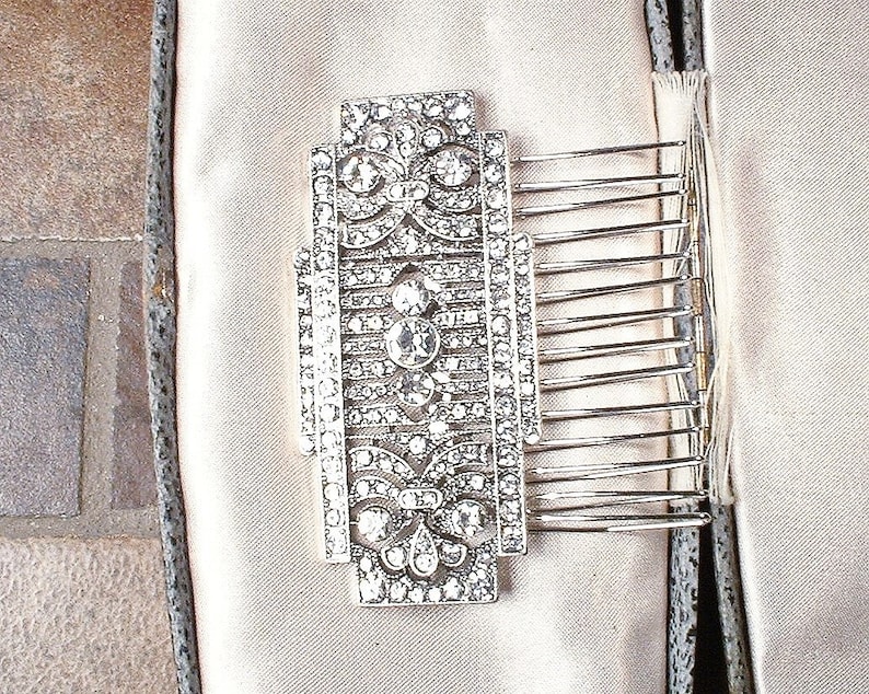 May include: A silver hair comb with a decorative design of clear rhinestones. The comb has a rectangular shape and is set on a white satin background.