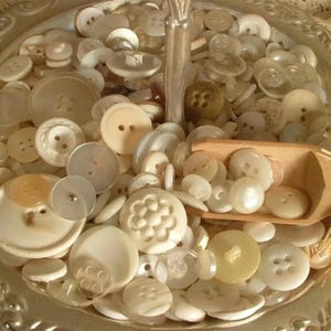 May include: A collection of white and cream coloured buttons of various sizes and shapes, some with a pearl-like finish, displayed in a silver bowl.
