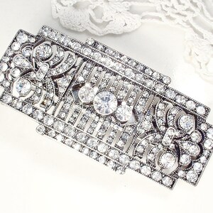 May include: A silver-toned, rectangular brooch with a geometric design. The brooch is adorned with clear rhinestones.