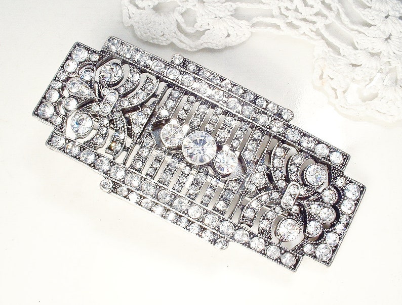 May include: A silver-toned, rectangular brooch with a geometric design. The brooch is adorned with clear rhinestones.