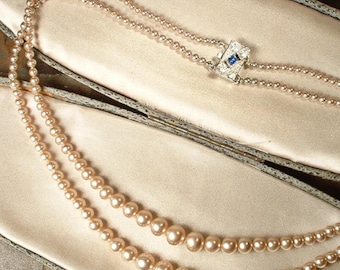 May include: A double strand pearl necklace with a silver clasp featuring a blue stone. The necklace is displayed in a beige velvet lined jewelry box.