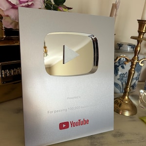 May include: A silver YouTube Play Button award, presented for passing 100,000 subscribers. The award features a reflective play button icon and the YouTube logo in red. The award is on a light-colored surface.