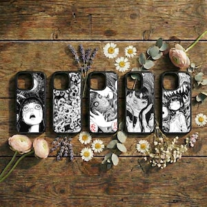 Puede incluir: Cinco fundas negras para teléfono con ilustraciones únicas en blanco y negro estilo manga. Las fundas están dispuestas sobre una superficie de madera rústica, rodeadas de flores y vegetación. Cada funda presenta un diseño diferente.