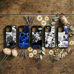 May include: Five black phone cases with unique designs, arranged on a wooden surface with flowers. The cases feature anime-style illustrations in black and white, and blue. One case has the text "BLUE LOCK".