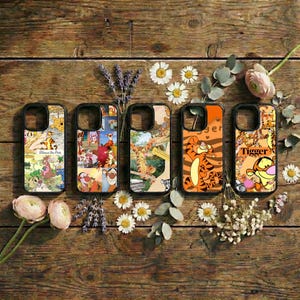 May include: Five phone cases with colorful cartoon designs featuring Winnie the Pooh characters. The cases are black with various images, including Tigger and other characters. The cases are arranged on a rustic wooden surface with flowers.