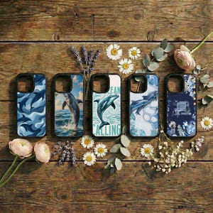 May include: Five phone cases with dolphin designs are displayed on a wooden surface. The cases feature various blue and white ocean-themed graphics, including leaping dolphins and wave patterns. One case has the text "Ocean Calling".