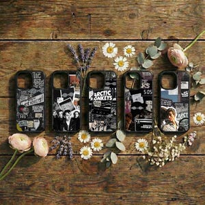 May include: Five black phone cases with Arctic Monkeys-themed collage designs. The cases feature black and white graphics, including text, photos, and the number "505". They are displayed on a wooden surface, decorated with flowers and foliage.