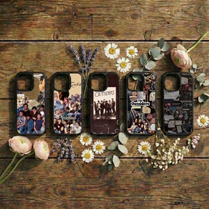 May include: Five phone cases with various designs, including collages and text. The cases feature images of people and text that reads "Gold" and "The Outsiders". The cases are arranged on a wooden surface with flowers.