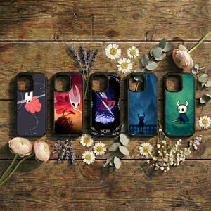 May include: Five black phone cases with unique designs featuring characters from a video game. The cases are arranged on a wooden surface with flowers and greenery. Each case has a different character and background.