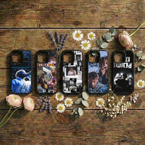 May include: Five black phone cases with various designs. One case features a blue and white image of a performer, another has a collage of images, and the others have portraits. The cases are arranged on a wooden surface with flowers.