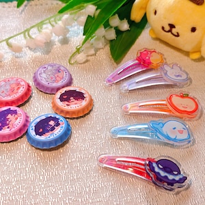 May include: A selection of vibrant hair clips and bottle cap magnets. The clips showcase designs like an apple, a bunny, and a smiling ghost. The bottle caps are decorated with anime-style character illustrations.