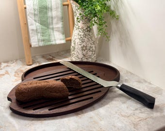 May include: A dark brown wooden bread cutting board with a loaf of bread and a serrated knife. The board has a slotted design for easy slicing. The knife has a black handle and a silver blade. A slice of bread is cut.
