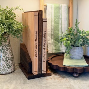 May include: Wooden cutting boards with labels for bread and cheese, meats and fish, and fruits and vegetables. The boards are in a dark wood holder. A decorative vase with greenery is on the left, and a wooden tray with a plant is on the right.