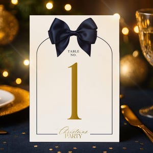 Può includere: Un biglietto con numero da tavolo color crema con un fiocco blu navy in alto. Il numero 1 è dorato, con le parole "Table No." sopra e "Christmas PARTY" sotto. Il biglietto è appoggiato su una tovaglia blu scuro.