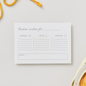 Puede incluir: Un bloc de notas blanco con una plantilla de rutina personalizada. La plantilla incluye secciones para tareas de la ma&ntilde;ana, la noche y peri&oacute;dicas, con l&iacute;neas para enumerar actividades. El bloc de notas tiene el texto "Custom routine for" en la parte superior y "Notes" en la parte inferior. Cuerda amarilla y un objeto decorativo en el fondo.