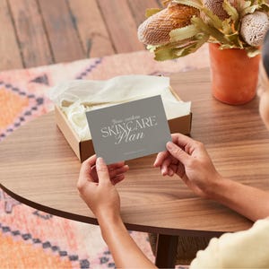Puede incluir: Una tarjeta gris con el texto "Your custom SKINCARE Plan" se sostiene sobre una mesa de madera. Una peque&ntilde;a caja con papel de seda se encuentra detr&aacute;s de la tarjeta. Un jarr&oacute;n con flores secas est&aacute; en el fondo.