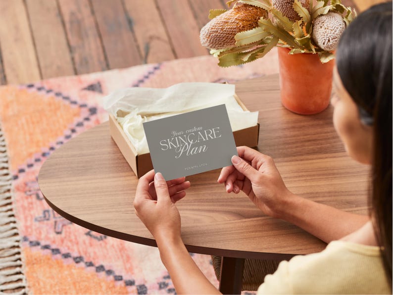 Puede incluir: Una tarjeta gris con el texto "Your custom SKINCARE Plan" se sostiene sobre una mesa de madera. Una peque&ntilde;a caja con papel de seda se encuentra detr&aacute;s de la tarjeta. Un jarr&oacute;n con flores secas est&aacute; en el fondo.