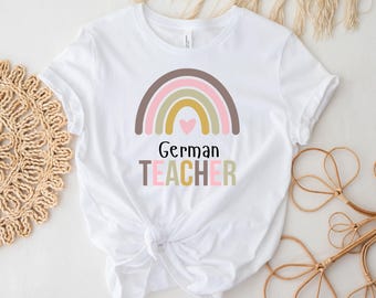 May include: White t-shirt featuring a rainbow design with a heart and the words "German TEACHER" in a mix of colors. The shirt is knotted at the waist and has short sleeves. The background includes decorative elements.