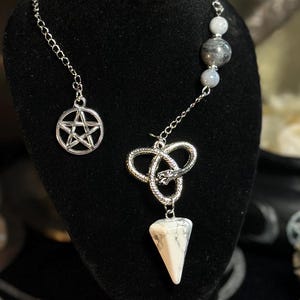 Op de afbeelding: Een zilverkleurige ketting met een pentagrambedel en een hanger met een witte en bruine steen. De ketting wordt getoond op een zwarte mannequinhoofd. De achtergrond bevat kristallen en een spiegel.