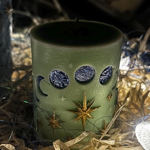May include: A sage green pillar candle featuring cut-out crescent moons and stars, filled with dark blue wax. Gold stars adorn the candle. The candle is surrounded by dried grass and fairy lights, with tarot cards in the background.