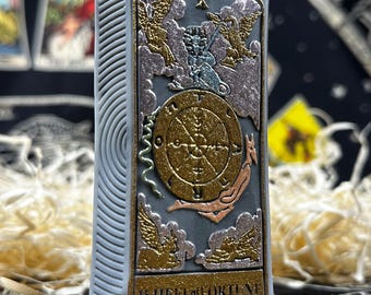May include: A rectangular, light gray candle with a detailed Wheel of Fortune tarot card design. The card features gold, copper, and silver accents, with the words "WHEEL OF FORTUNE" at the bottom. The candle has a textured side.