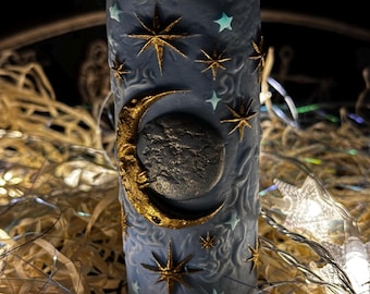 May include: A cylindrical candle with a celestial design. The candle is a muted blue color, adorned with gold stars, a crescent moon, and a textured, dark gray moon. The design is embossed on the candle's surface, creating a three-dimensional effect. The candle is surrounded by decorative lights.