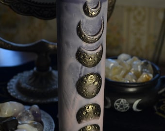May include: A lavender pillar candle with gold crescent moon accents. The candle features a series of embossed moon phases. The candle is cylindrical and has a wick at the top. The candle is a decorative item.