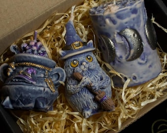 May include: A box containing three blue candles. One is a cauldron, another is a cat wearing a witch hat and holding a broom, and the third is a pillar candle with crescent moon accents. The candles are surrounded by straw.