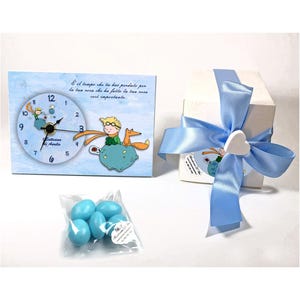 Può includere: Un orologio azzurro con un'illustrazione a cartoni animati di un ragazzo e una volpe, accanto a una scatola regalo bianca legata con un nastro blu e un ornamento a forma di cuore. È presente anche un sacchetto di caramelle blu.