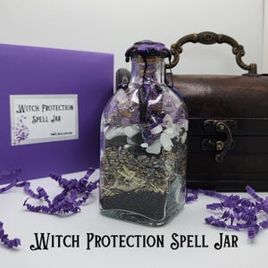 May include: A clear glass jar filled with layers of ingredients, including black sand, herbs, and crystals. The jar is sealed with a cork and purple wax. A purple box and a wooden chest are in the background. The text "Witch Protection Spell Jar" is visible.