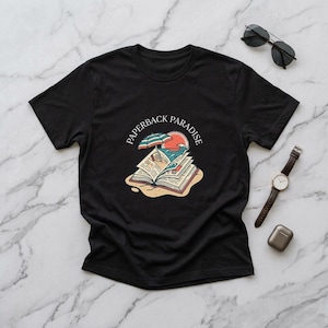 May include: A black short-sleeved t-shirt featuring a graphic of an open book with a beach umbrella, ocean waves, and a sun. The text PAPERBACK PARADISE is printed in an arc above the illustration. The shirt is displayed with sunglasses and a watch.