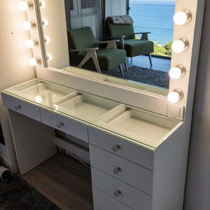 Puede incluir: Tocador blanco con un gran espejo rectangular enmarcado con bombillas. El tocador tiene una tapa de cristal con compartimentos de almacenamiento y cajones. El espejo refleja una vista del mar y dos sillas verdes.