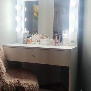 K&ouml;nnte beinhalten: Wei&szlig;er Schminktisch mit einem gro&szlig;en Spiegel, der von runden Lichtern umrahmt ist. Der Tisch hat eine Schublade und einen Stuhl mit rosa Kunstfell. Make-up und Accessoires sind ausgestellt.