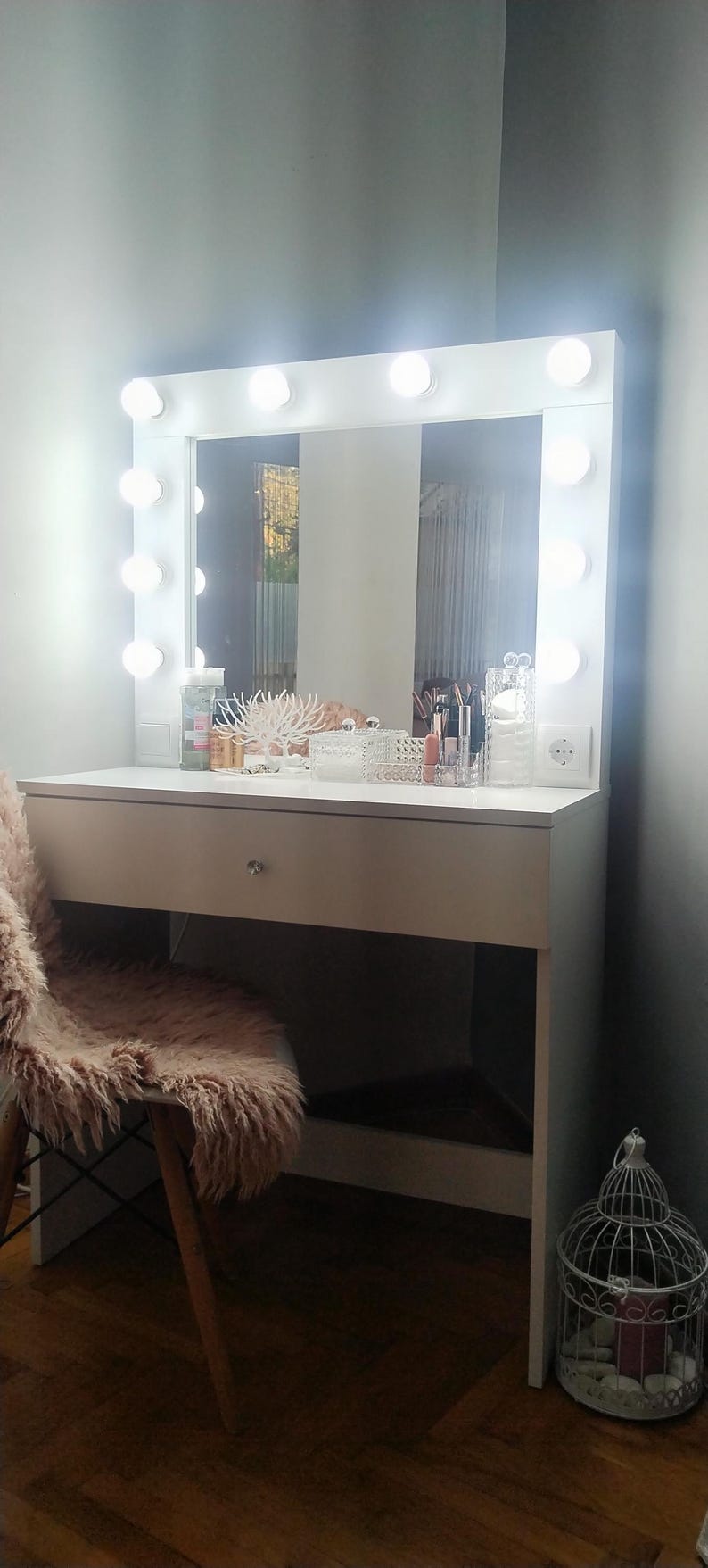 K&ouml;nnte beinhalten: Wei&szlig;er Schminktisch mit einem gro&szlig;en Spiegel, der von runden Lichtern umrahmt ist. Der Tisch hat eine Schublade und einen Stuhl mit rosa Kunstfell. Make-up und Accessoires sind ausgestellt.
