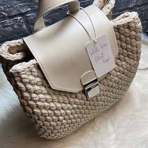 May include: A beige crocheted handbag featuring a smooth cream-colored flap with a silver-toned metal clasp. A white rectangular tag is attached to the handle, displaying the handwritten text "With love, Glori bags." The bag has a structured base.