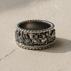 Può includere: Un anello in metallo color argento con un dettagliato rilievo scultoreo dell'Ultima Cena lungo la fascia esterna. L'anello ha una forma cilindrica spessa con una texture a corda lungo i bordi superiore e inferiore, raffigurante varie figure.