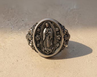 May include: A silver-toned ring featuring an oval face with a raised relief of the Virgin of Guadalupe surrounded by detailed rose motifs. The band and sides of the ring are decorated with intricate floral carvings and textured metalwork.