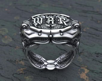 May include: A silver ring with a detailed design featuring a skull on each side and the word "WAR" in gothic lettering on the top.