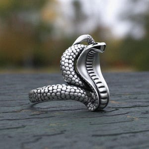 Può includere: Un anello color argento a forma di serpente cobra. La testa del serpente è sollevata, con il cappuccio aperto e la bocca aperta. L'anello ha un motivo strutturato a forma di scaglia.