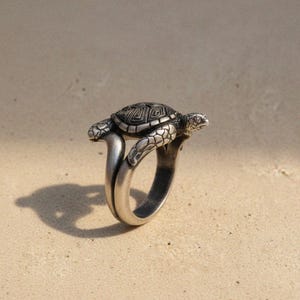 Op de afbeelding: Een zilverkleurige ring met een gedetailleerd schildpadontwerp. De schildpad heeft een geometrisch patroon op de schild, en de ringband buigt omhoog om de schildpad te ondersteunen. De ring staat tegen een neutrale achtergrond.