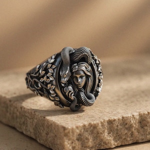 May include: A silver-toned ring featuring a detailed design of a woman's face intertwined with snakes. The ring has an oval face with intricate leaf and vine detailing around the edges. The ring is displayed on a textured, light brown surface.
