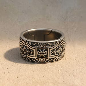 Puede incluir: Un anillo plateado con un diseño intrincado que presenta una cruz de Malta y motivos florales. El anillo tiene una banda ancha y una superficie detallada en relieve. El anillo es probablemente de metal y se muestra sobre una superficie neutra.
